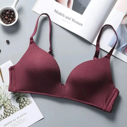 A maroon wireless bra with molded cups and adjustable spaghetti straps, resting on a gray surface next to a magazine and a small bowl of coffee beans.