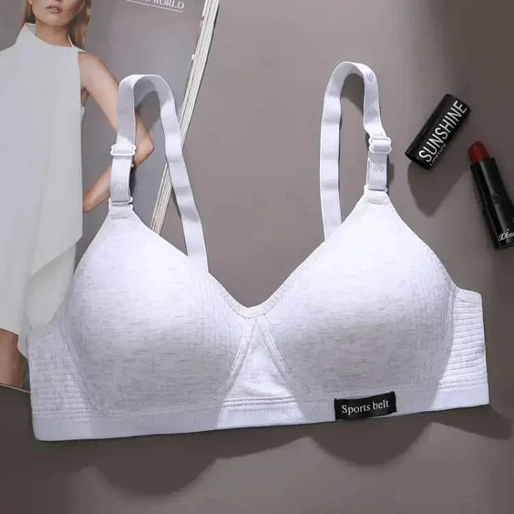 A light grey wireless sports bra with wide adjustable straps and a small black tab labeled Sports belt at the bottom sits on a grey surface next to a magazine and two lipsticks.