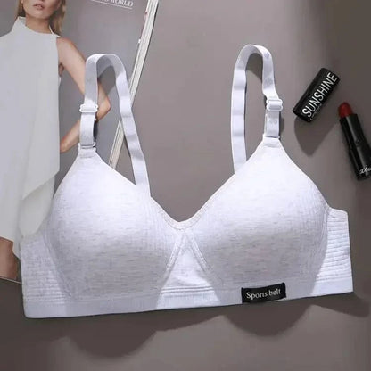 A light grey wireless sports bra with wide adjustable straps and a small black tab labeled Sports belt at the bottom sits on a grey surface next to a magazine and two lipsticks.