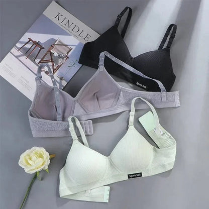 Three soft bras are displayed on a grey surface, featuring black, grey, and light green colors with ribbed fabric and adjustable straps, accompanied by a white artificial rose and a book in the background.