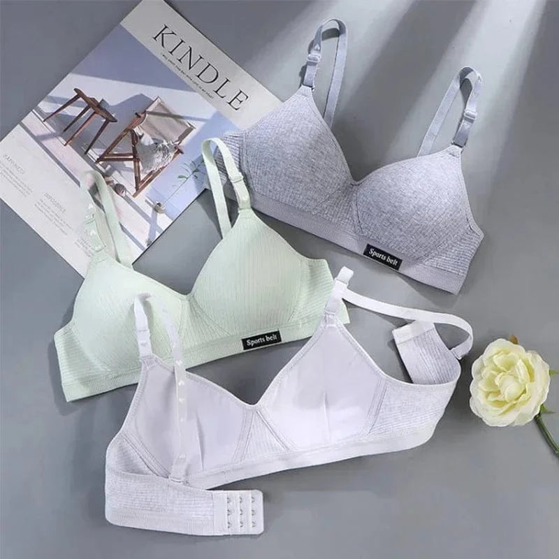 Three soft, wireless bras in grey, light green, and white are arranged on a grey surface next to a magazine and a white artificial flower.