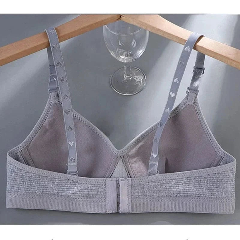A grey wireless bra with adjustable straps featuring a subtle heart pattern, shown hanging on a wooden hanger above a glass.