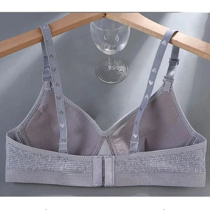 A grey wireless bra with adjustable straps featuring a subtle heart pattern, shown hanging on a wooden hanger above a glass.