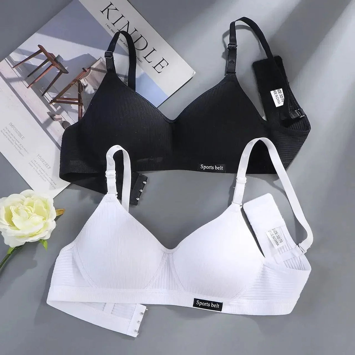 Two wireless bras with thin adjustable straps and small rectangular Sports belt labels, one in black and one in white, are displayed on a grey surface next to a flower and a Kindle advertisement.
