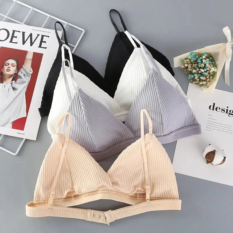 Three ribbed triangle bras in black, white, and beige are laid out on a gray surface next to a magazine and a decorative dried flower bouquet.