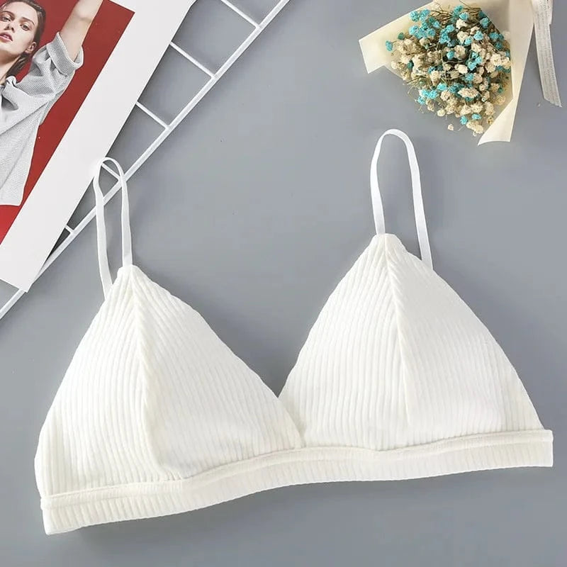 A white ribbed triangle bralette with thin adjustable straps lies on a grey surface next to a small bouquet of dried flowers and a magazine.