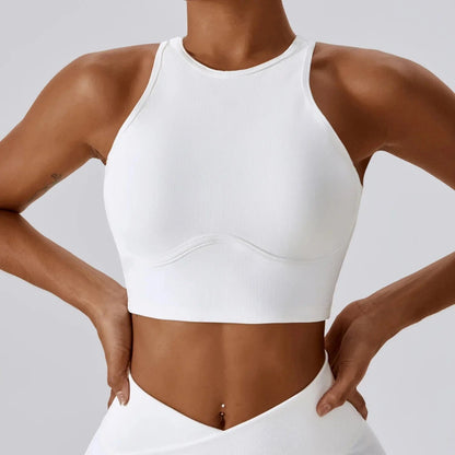 A white sleeveless athletic crop top with a high neckline and a curved seam detail under the bust, worn by a person with their hands on their hips.