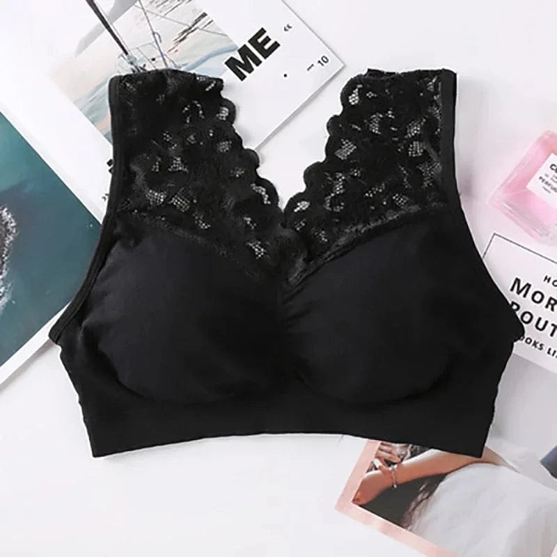A black wireless bralette with a deep V-neckline and decorative floral lace trim along the straps and front edges, displayed flat on a white surface with magazines and a small bottle of perfume.