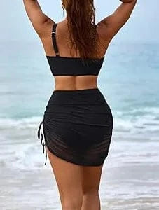 A black two-piece swimsuit with a ruched drawstring side-tie skirt worn by a woman standing on a beach with the ocean in the background.