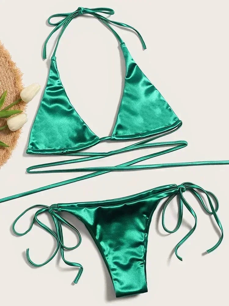 A vibrant green satin bikini set featuring a triangle top with long wrap-around ties and matching side-tie bikini bottoms laid flat on a neutral background with a straw hat and tulips nearby.