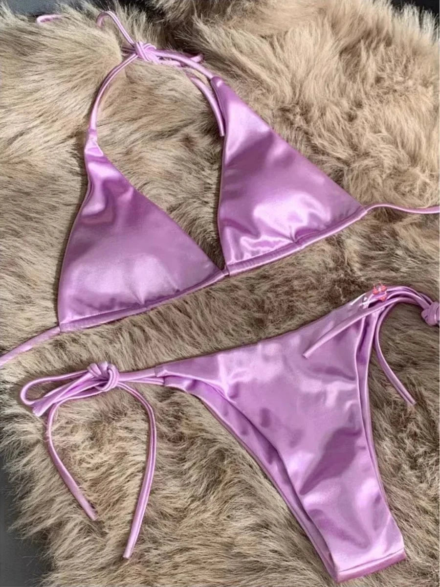 A light purple satin two-piece bikini set with a triangle top and tie-side bottoms, displayed against a textured faux fur background.